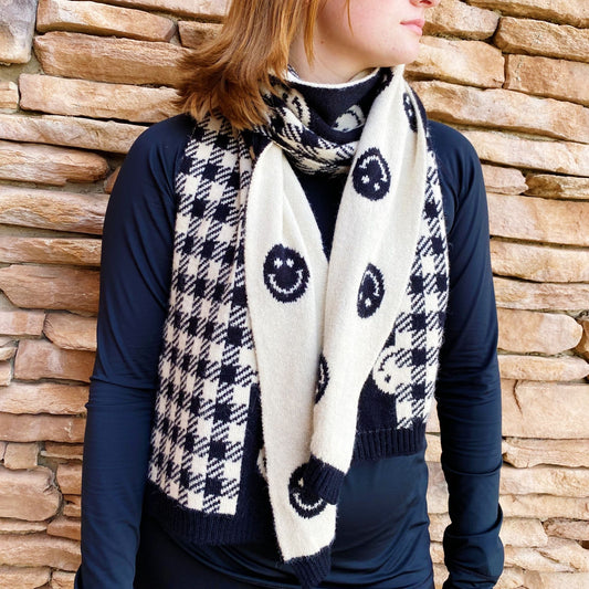 Stylish black and white patterned scarf featuring smiley faces, worn by a woman against a stone wall backdrop, perfect for adding a cheerful touch to any outfit.