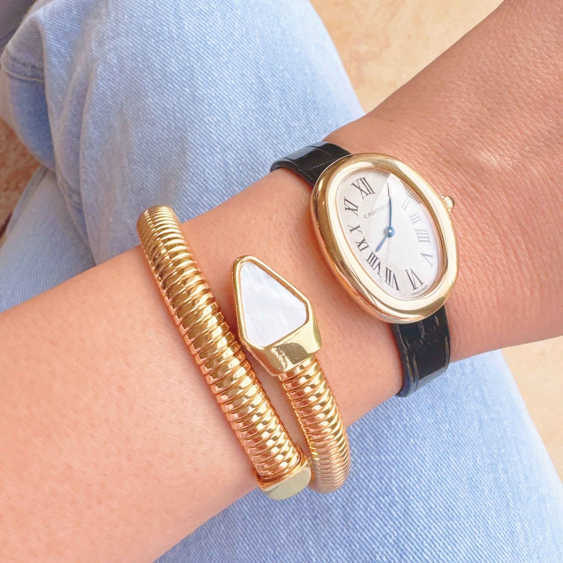 A close-up of a woman's wrist wearing a stylish gold-tone bracelet and an elegant oval-shaped Cartier watch with a black leather strap, set against a backdrop of light blue denim.