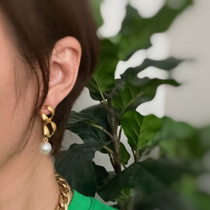 Elegant gold earrings with twisting design and pearl accent, worn by a woman against a backdrop of green foliage.