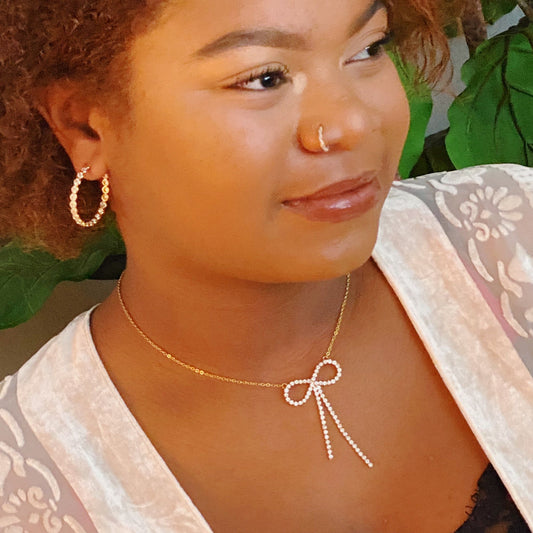 Elegant gold necklace featuring a bow design adorned with sparkling rhinestones, paired with matching hoop earrings, against a backdrop of lush green leaves.