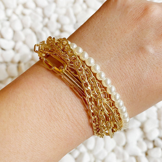 Gold layered bracelets with pearl accents worn on a wrist, against a textured white stone background.