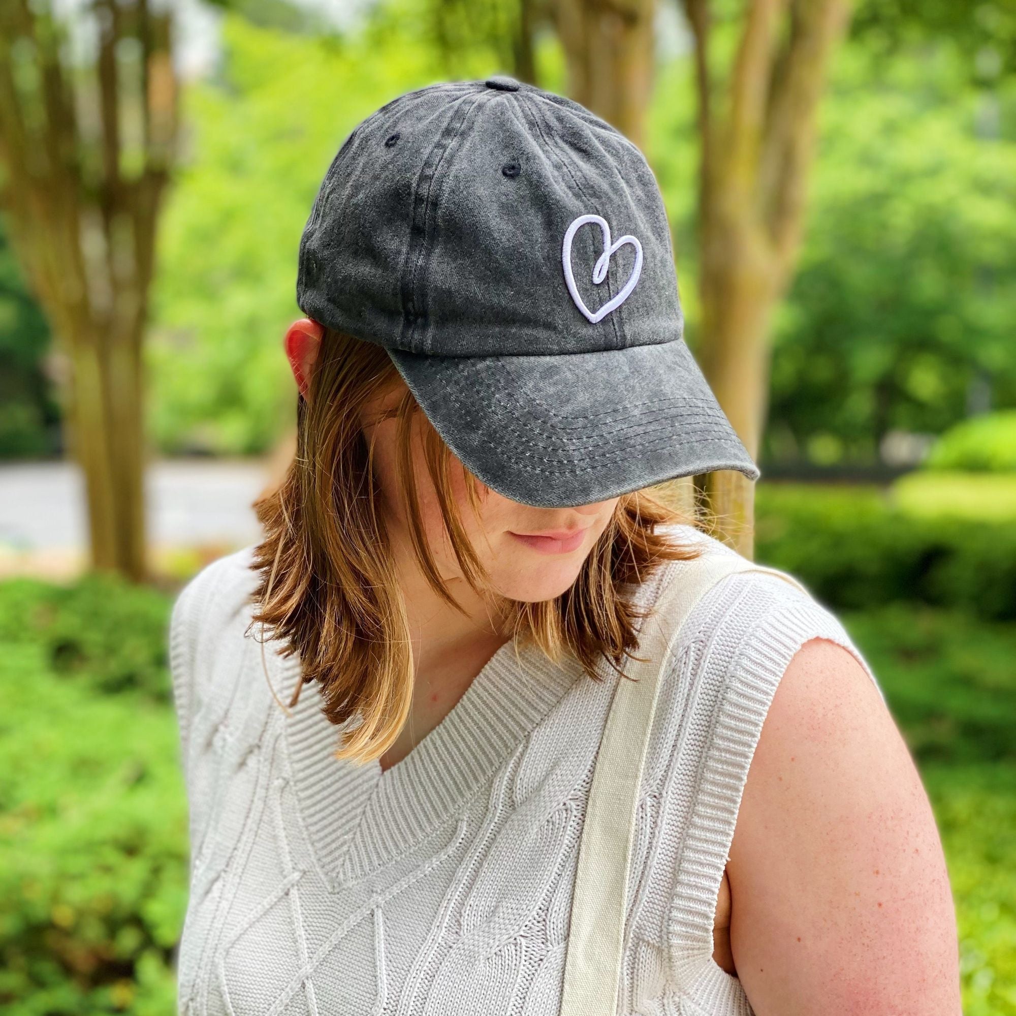 Casual gray baseball cap featuring a white heart embroidery, worn by a woman outdoors, surrounded by lush green foliage.