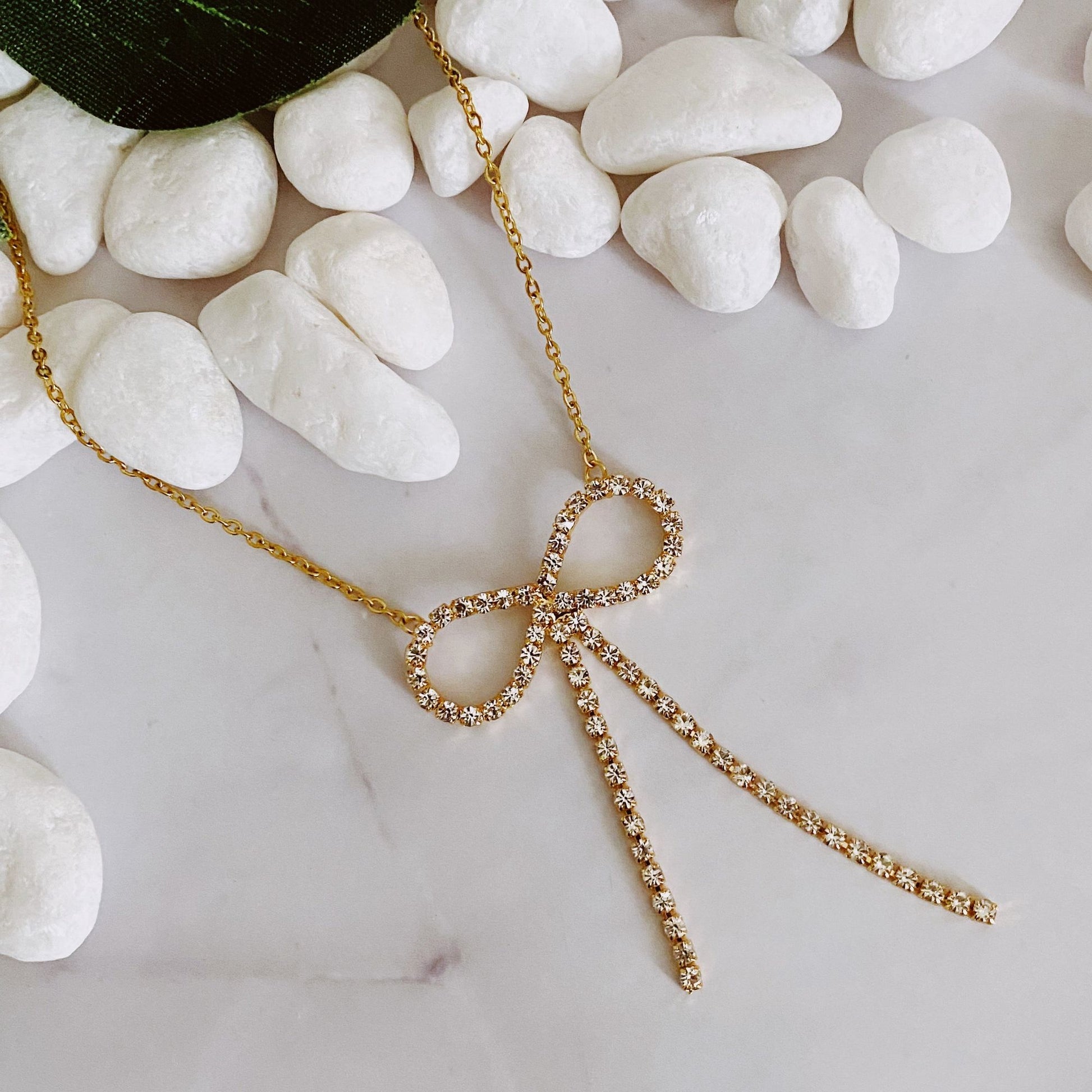 Gold necklace with a sparkling rhinestone bow pendant resting on white pebbles against a marble background.