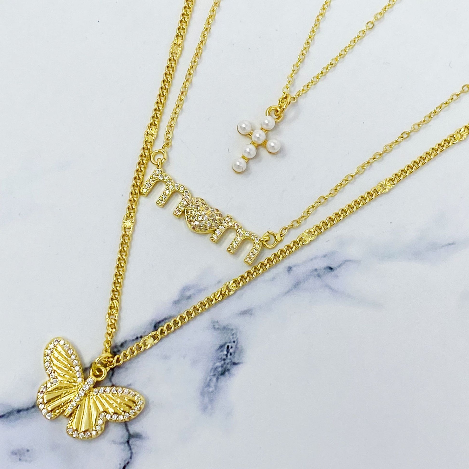 Gold layered necklace set featuring a butterfly pendant, a sparkling "mom" charm with a heart, and a delicate pearl cross charm, all on marble background.