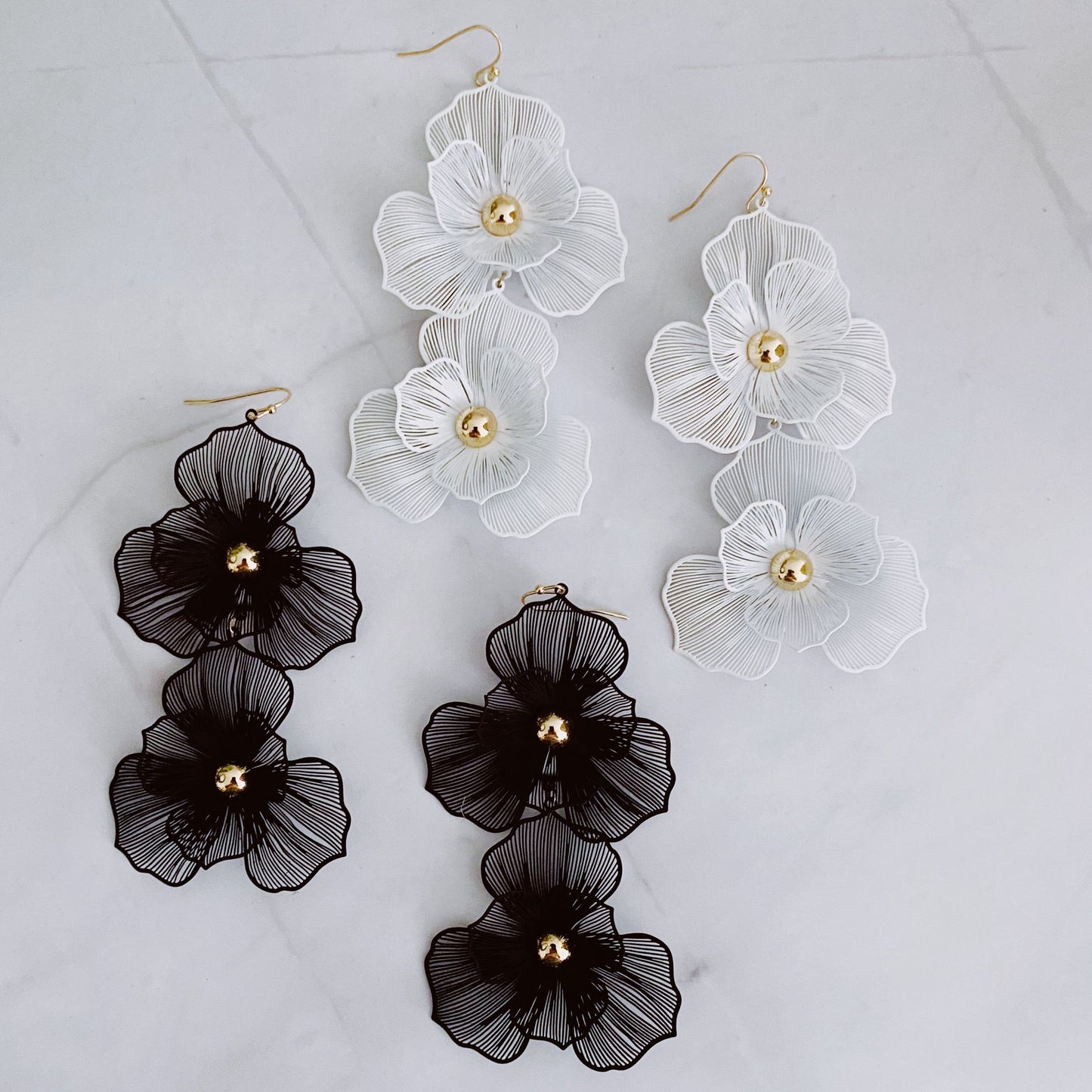 Chic statement earrings featuring three-dimensional floral designs in black and white, embellished with gold accents, displayed on a marble background.
