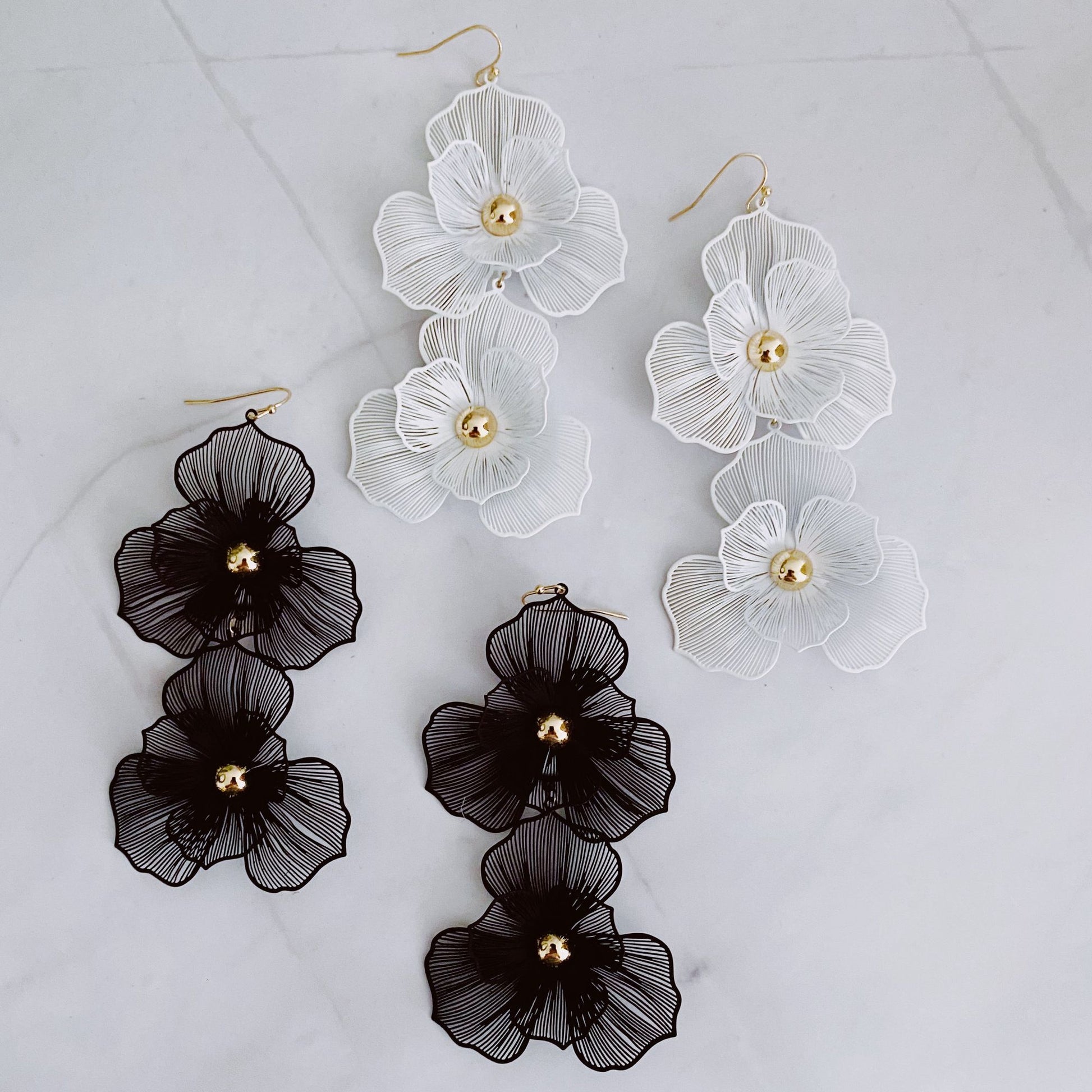 Chic statement earrings featuring three-dimensional floral designs in black and white, embellished with gold accents, displayed on a marble background.