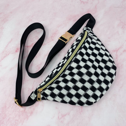 Black and white checkered fanny pack with a gold zipper and adjustable black strap on a pink marble background.