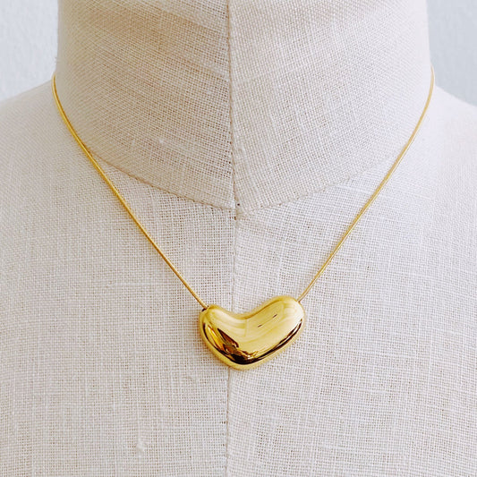 Gold heart-shaped pendant necklace displayed on a beige fabric mannequin, showcasing its glossy finish and elegant design.