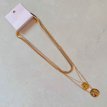 Gold layered necklace with unique charms on a display card, resting on a light-colored textured surface.