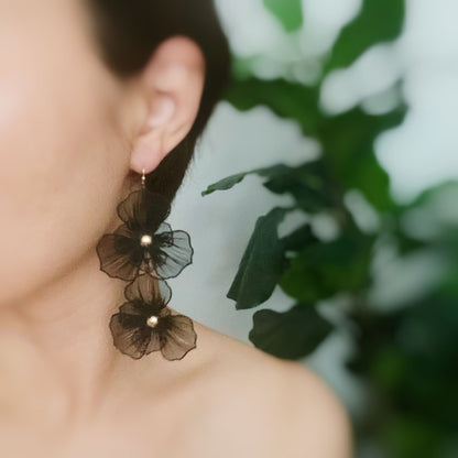 Elegant black floral earrings with pearl accents, worn by a woman with a simplistic background featuring green foliage.