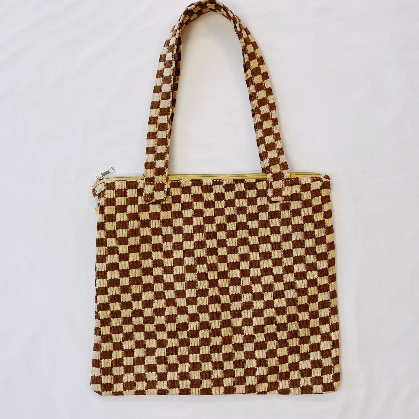 Brown and cream checkered tote bag with handles, featuring a yellow zipper, displayed on white background.