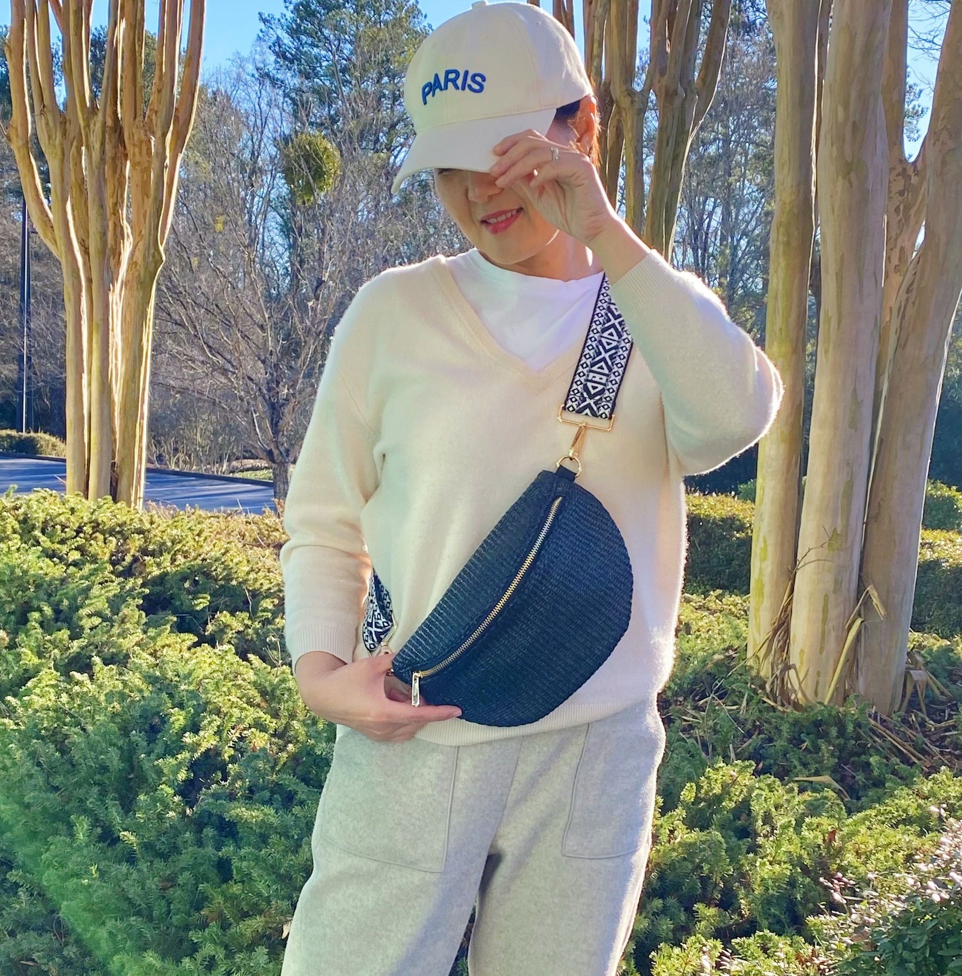 Stylish woman wearing a light sweater and gray sweatpants, holding a trendy black crossbody bag with a patterned strap, set against a scenic outdoor background with greenery and trees.