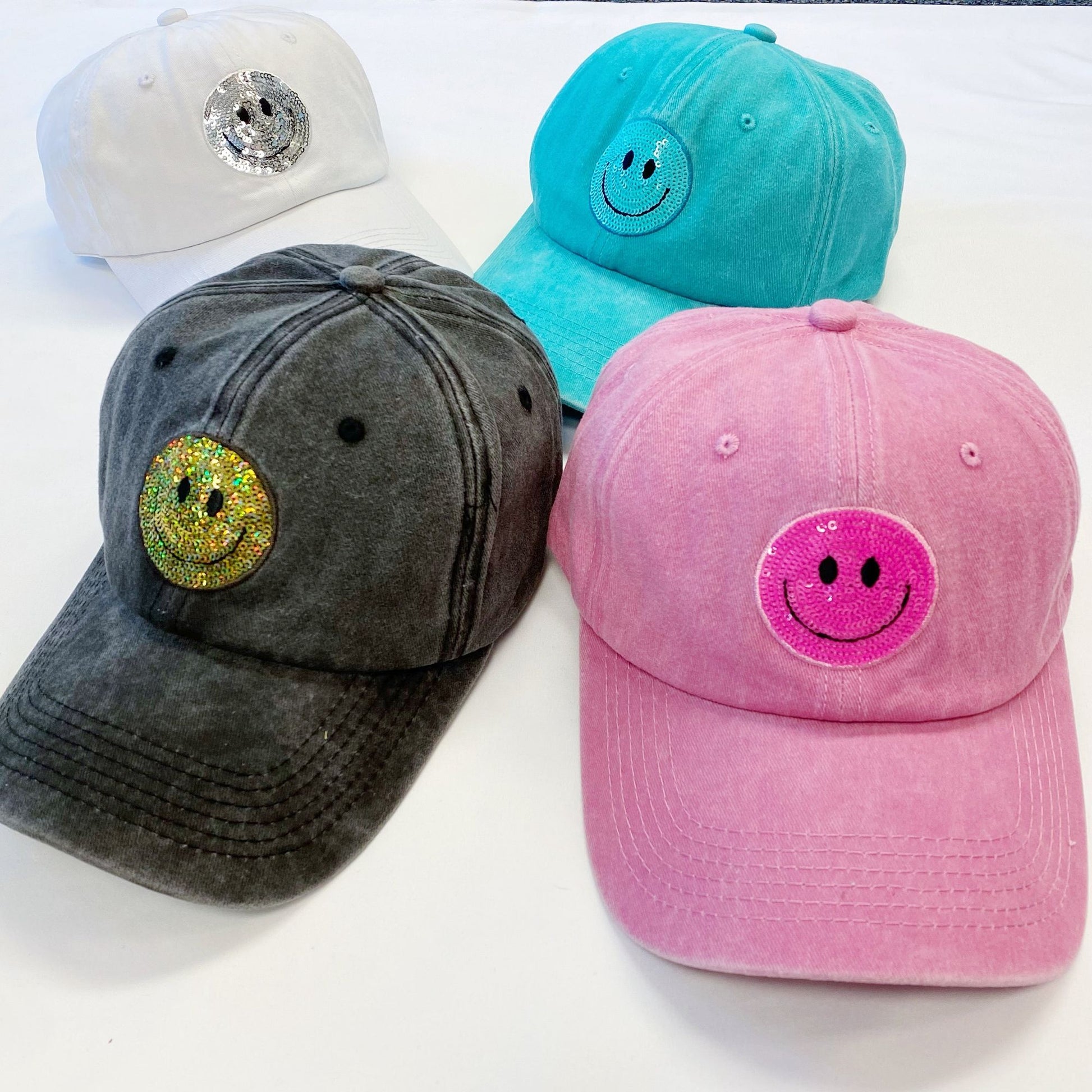 Four trendy cotton caps featuring colorful smiley face patches, including white, turquoise, gray with sparkly yellow smiley, and light pink varieties, displayed against a plain white background.