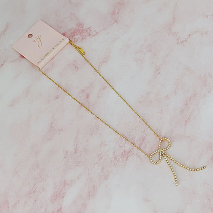 Gold necklace with a sparkling rhinestone bow pendant, elegantly displayed on a pale pink marble background.