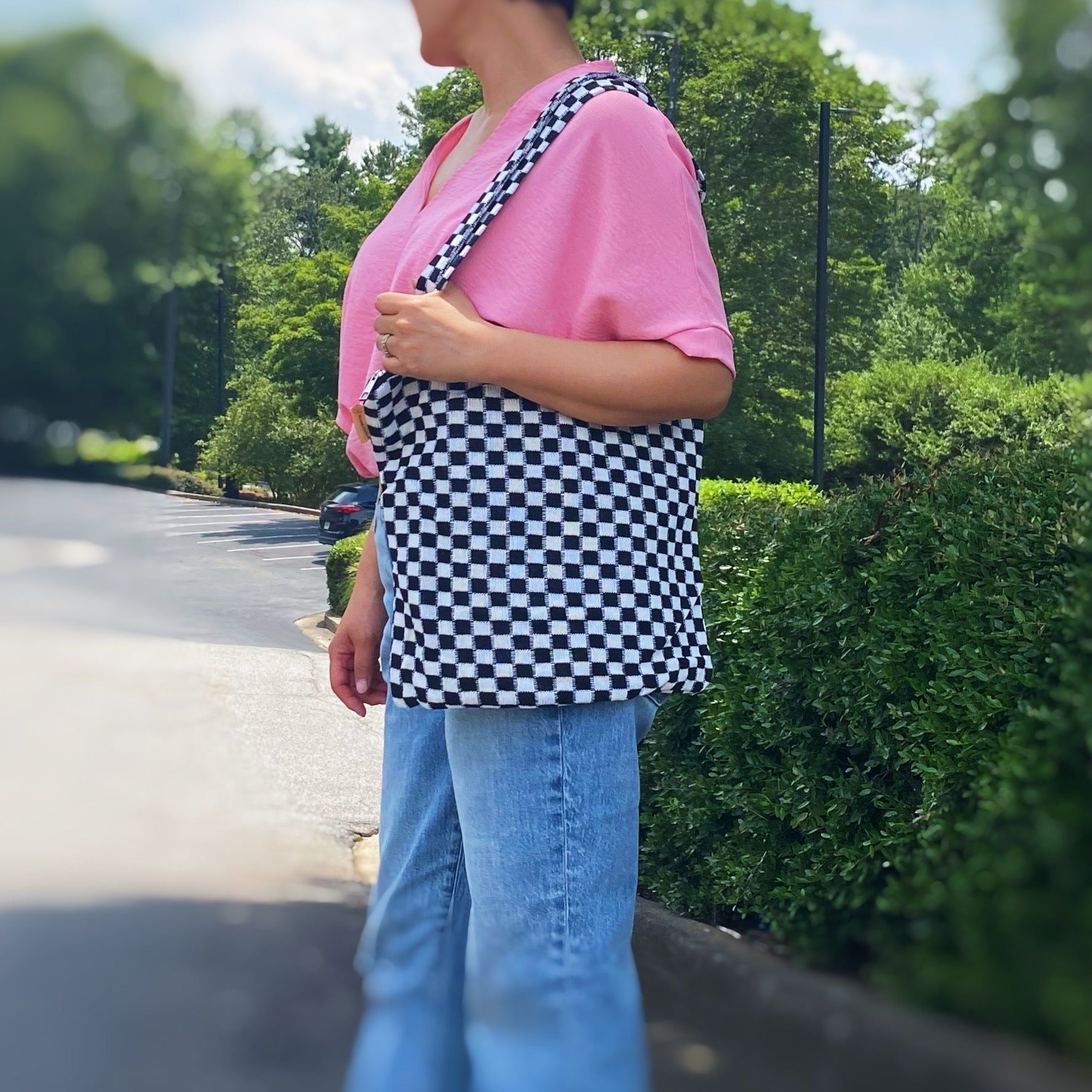 Woman wearing a pink top and blue jeans, holding a stylish black and white checkered tote bag while standing outdoors in a sunny setting with greenery in the background.