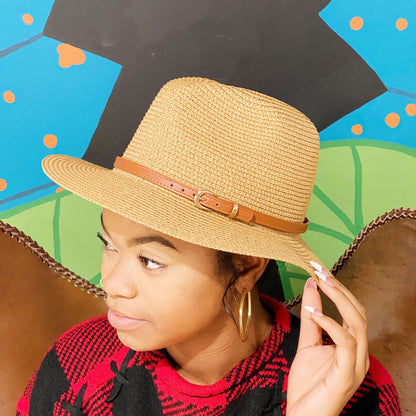 Stylish straw fedora hat with a brown leather band, worn by a person with curly hair, against a colorful mural background.