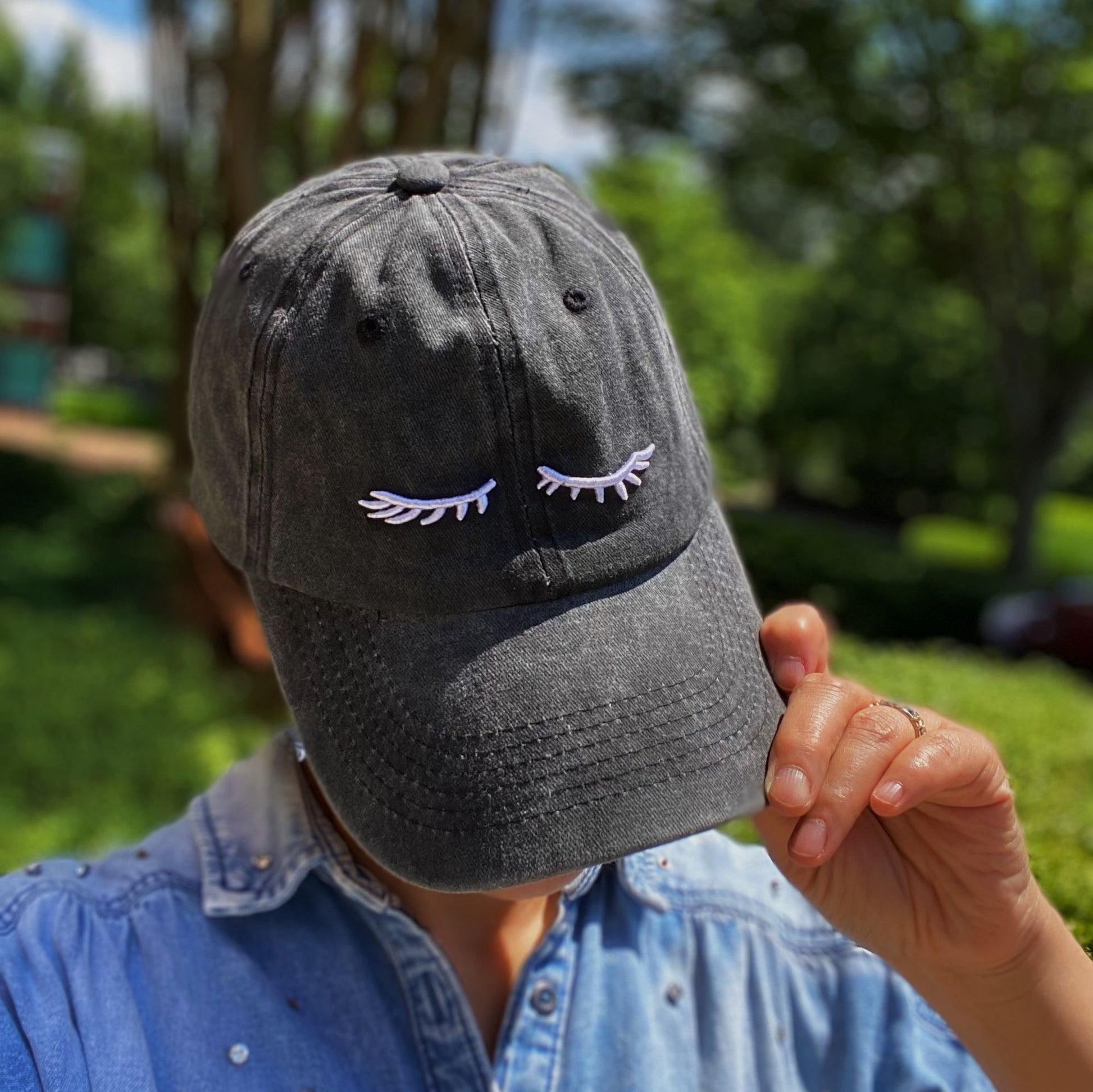 Gray baseball cap with white embroidered eyelash design, worn by a person in a denim shirt against a lush green background.