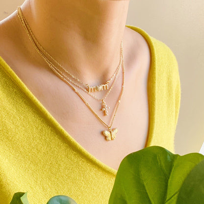 Layered gold necklaces featuring a 'mom' pendant, pearl accents, and a butterfly charm, worn by a model in a bright yellow sweater, complemented by green foliage.