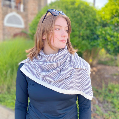 Woman wearing a stylish gray and white herringbone-patterned scarf over a black long-sleeve shirt, set against a lush green background.