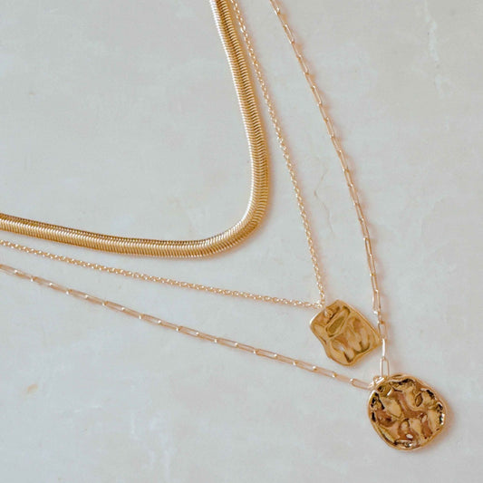 Three-layered gold necklace featuring a textured pendant, flat chain, and flexible snake chain on a light marble background.