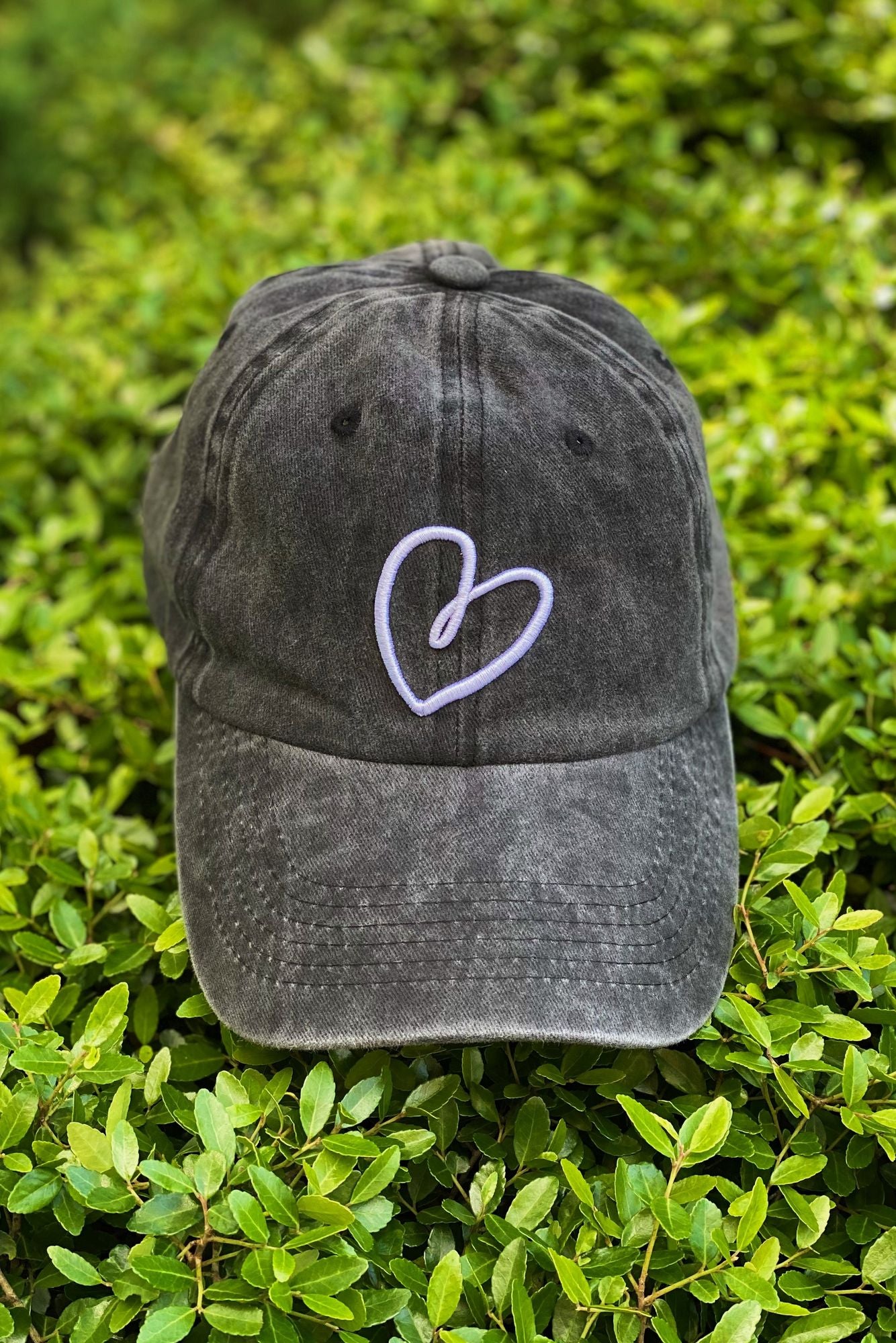 Dark gray cap with a white heart embroidered on the front, resting on a bed of vibrant green leaves.