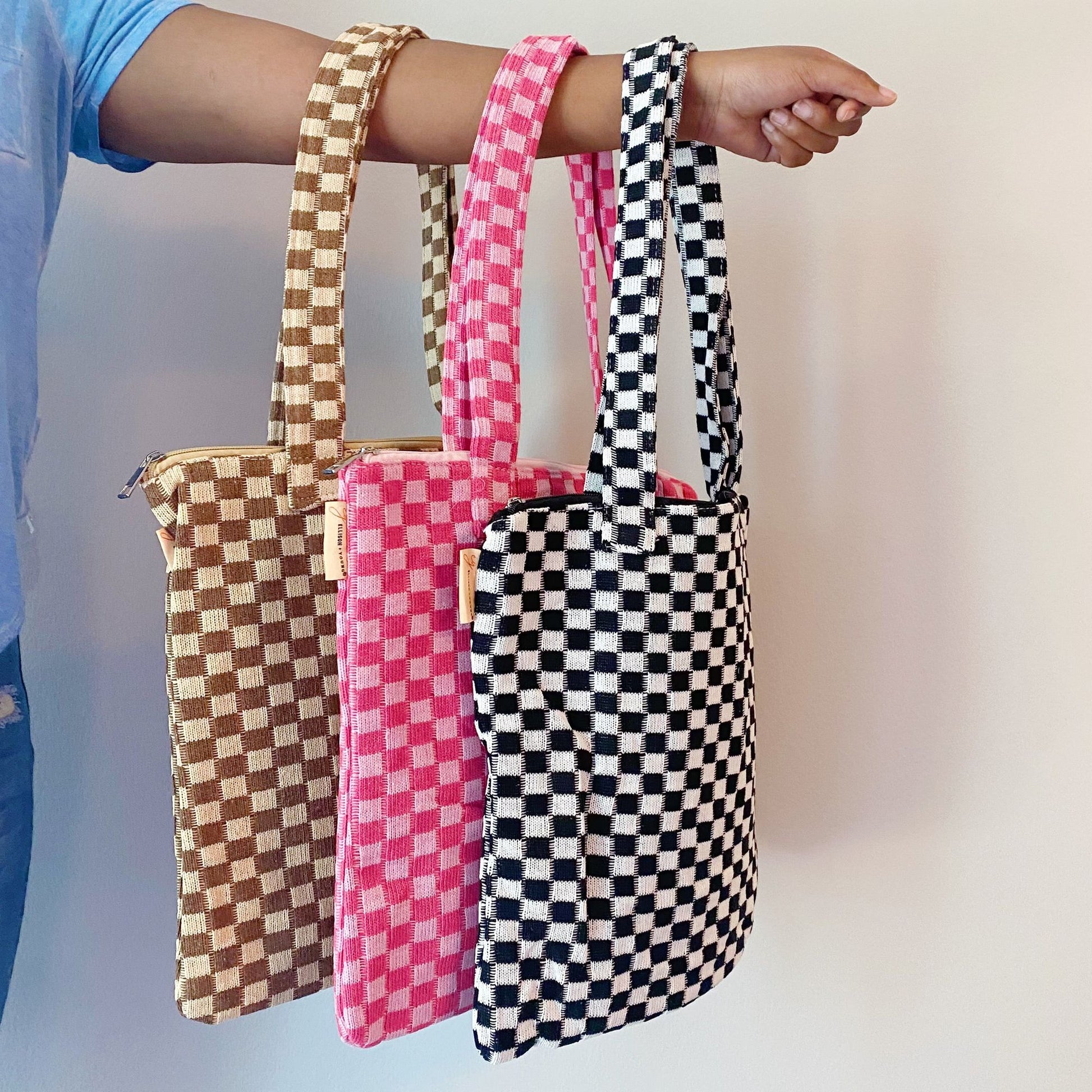 A hand holding three stylish tote bags in checkered patterns—brown, pink, and black—showcasing their unique designs and sturdy handles against a plain background.