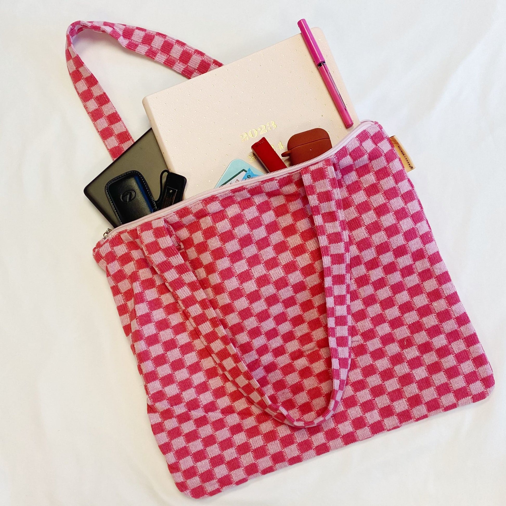 Pink gingham patterned tote bag filled with a planner, pens, keys, and cards against a white background.