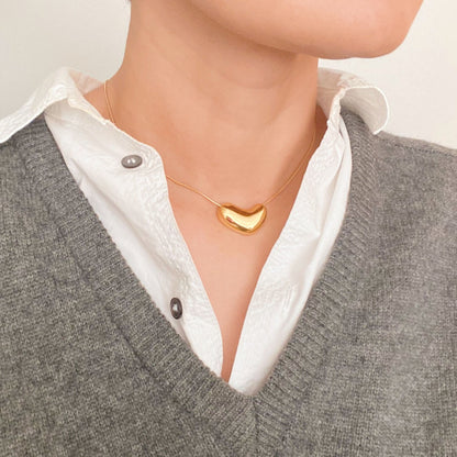 Stylish gold heart-shaped pendant necklace worn over a white button-up shirt, paired with a grey sweater, showcasing contemporary jewelry fashion.