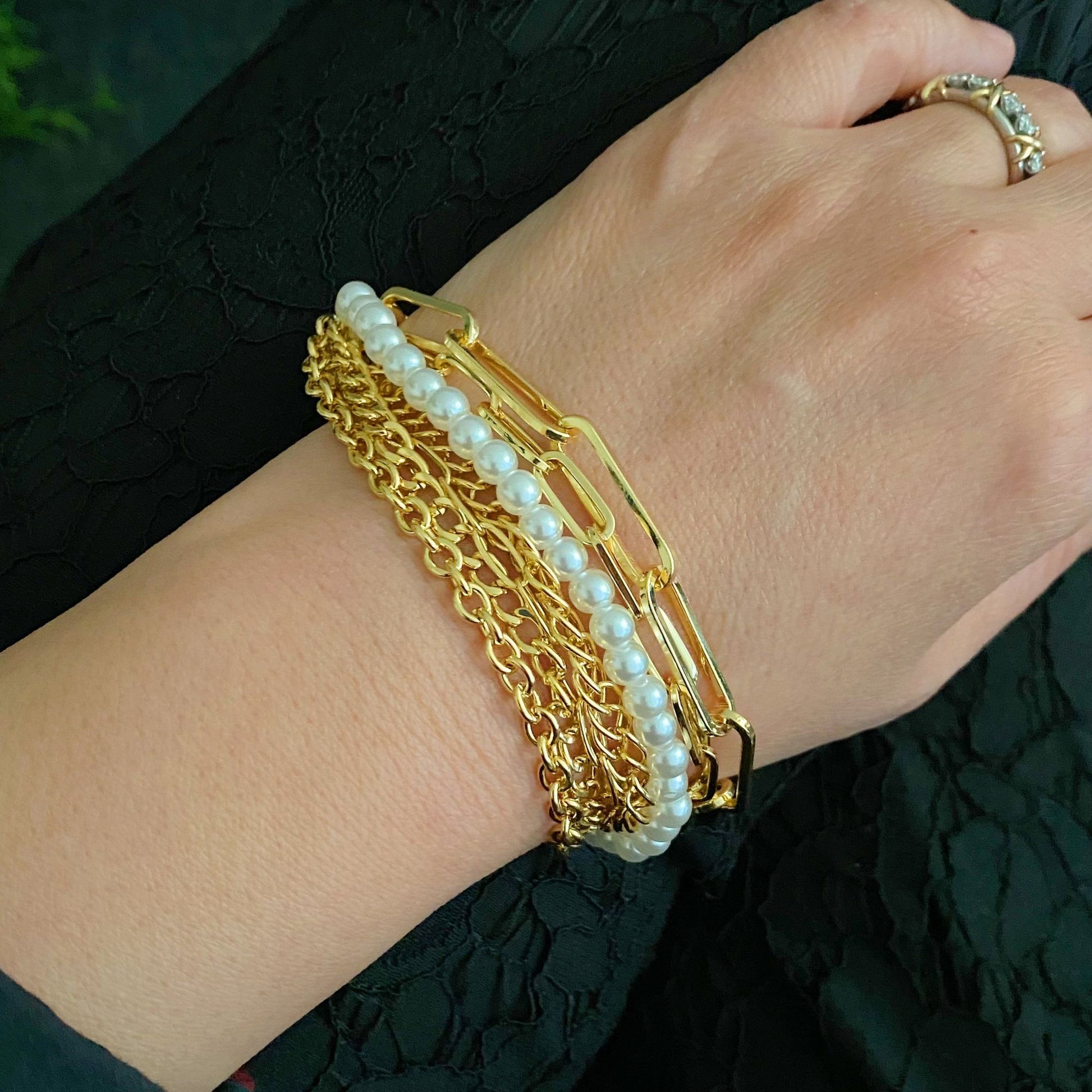 Gold and pearl layered bracelet worn on a woman's wrist, showcasing unique chain links and elegant pearls against a black textured background.