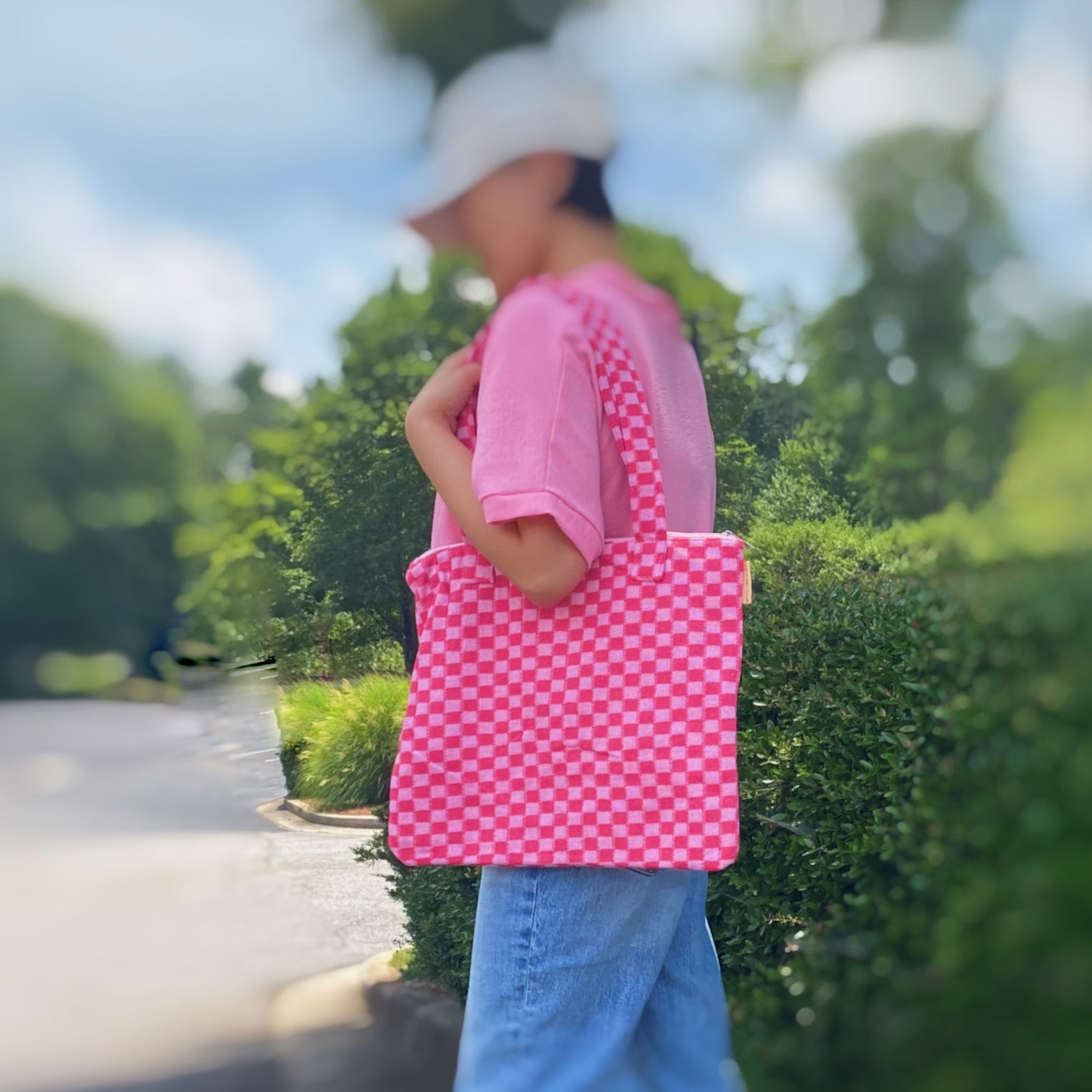 A woman wearing a pink shirt and a white cap holds a vibrant pink and white checkered tote bag against a blurred outdoor background with greenery.