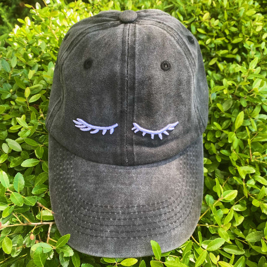 Black baseball cap with embroidered eyelashes design resting on lush green foliage.