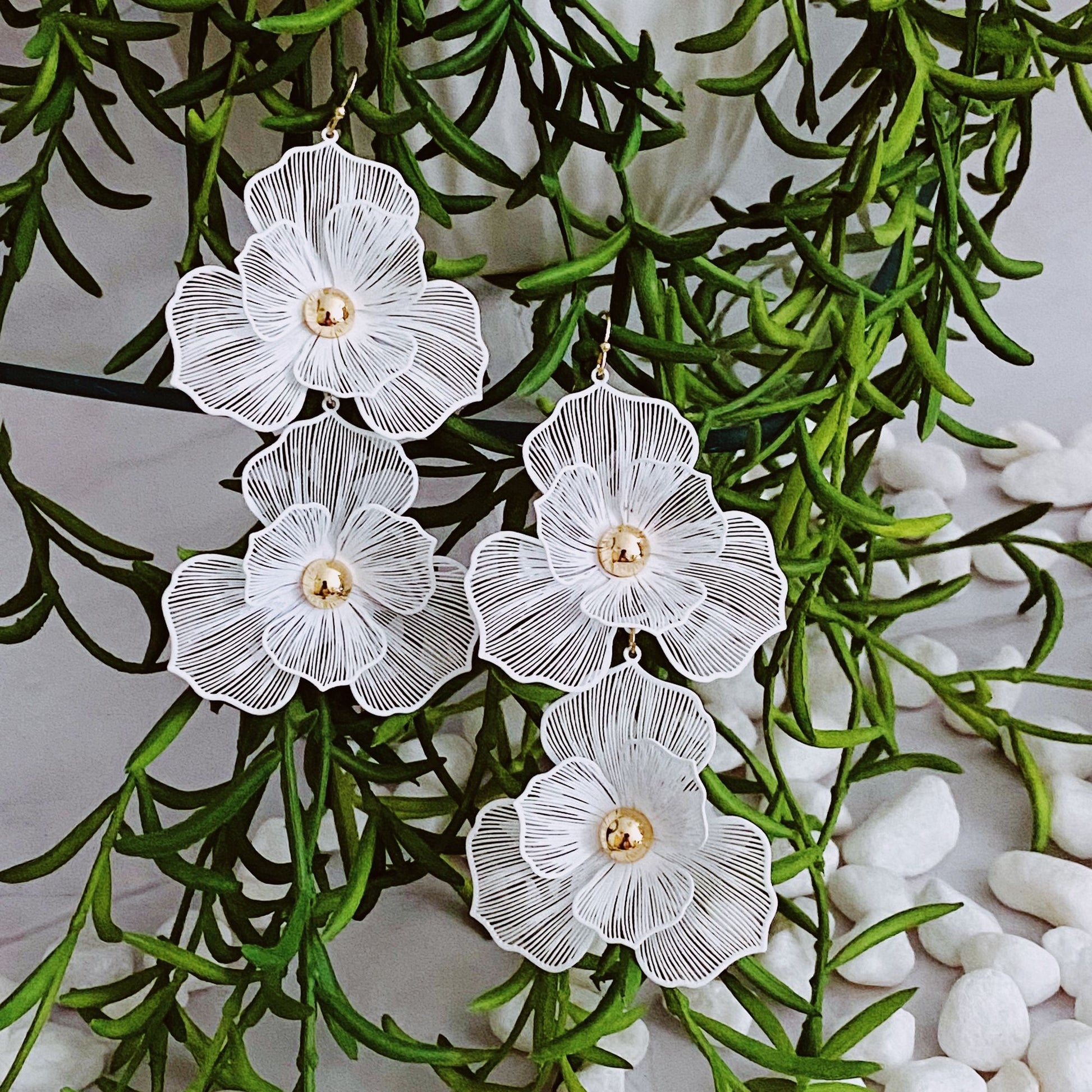 Elegant white floral earrings featuring intricate lace-like details and gold center, hanging among lush greenery and decorative pebbles, perfect for adding a touch of sophistication to any outfit.
