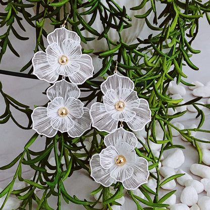 Elegant white floral earrings featuring intricate lace-like details and gold center, hanging among lush greenery and decorative pebbles, perfect for adding a touch of sophistication to any outfit.