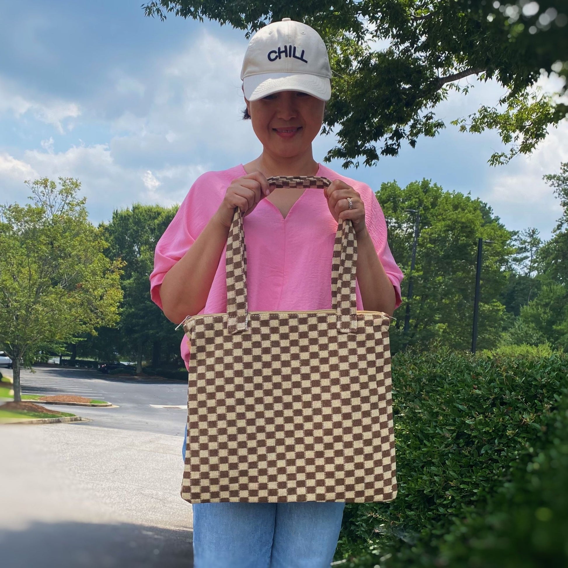 Stylish brown and cream checkered tote bag held by a woman wearing a pink shirt and a chill cap, set against a natural outdoor backdrop with greenery and a partially cloudy sky.
