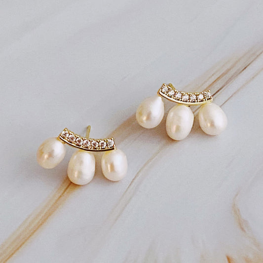 Elegant gold and pearl earrings featuring a curved design adorned with sparkling rhinestones and three dangling freshwater pearls, set against a soft marble background.