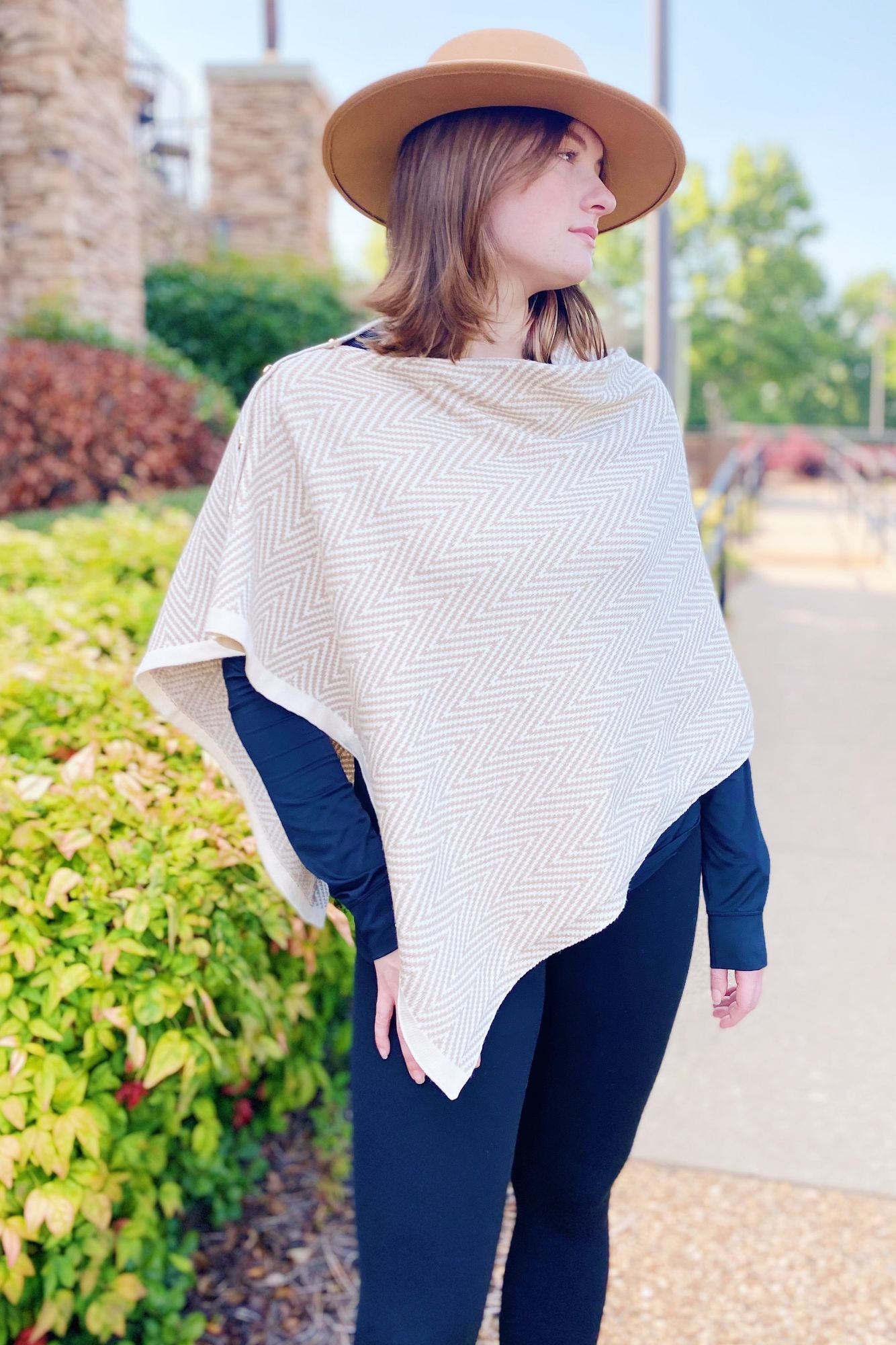 Fashionable lightweight beige herringbone poncho worn over a black top, paired with a wide-brim hat, surrounded by greenery in an outdoor setting.