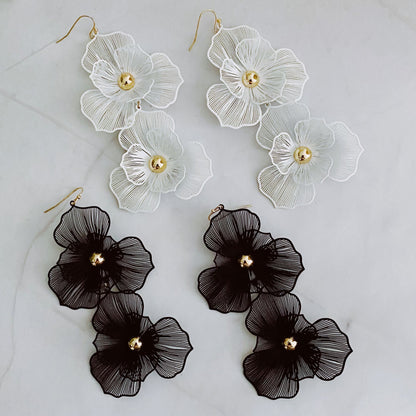 Elegant floral earrings in white and black, featuring intricate layered petal designs with a central gold bead, displayed on a marble background.