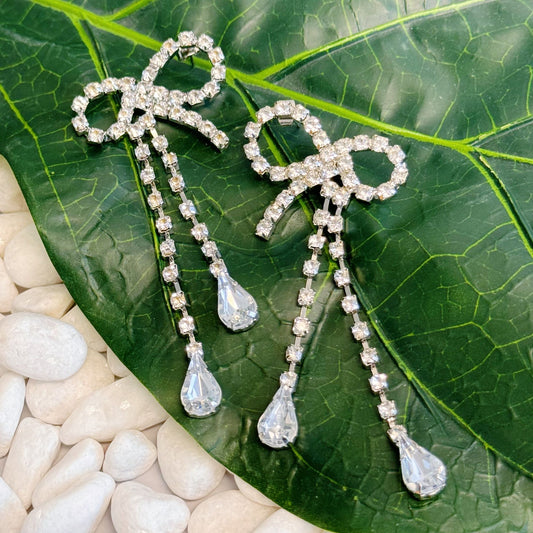 Elegant crystal bow earrings with dangling teardrop gemstones, displayed on a green leaf background surrounded by white pebbles.