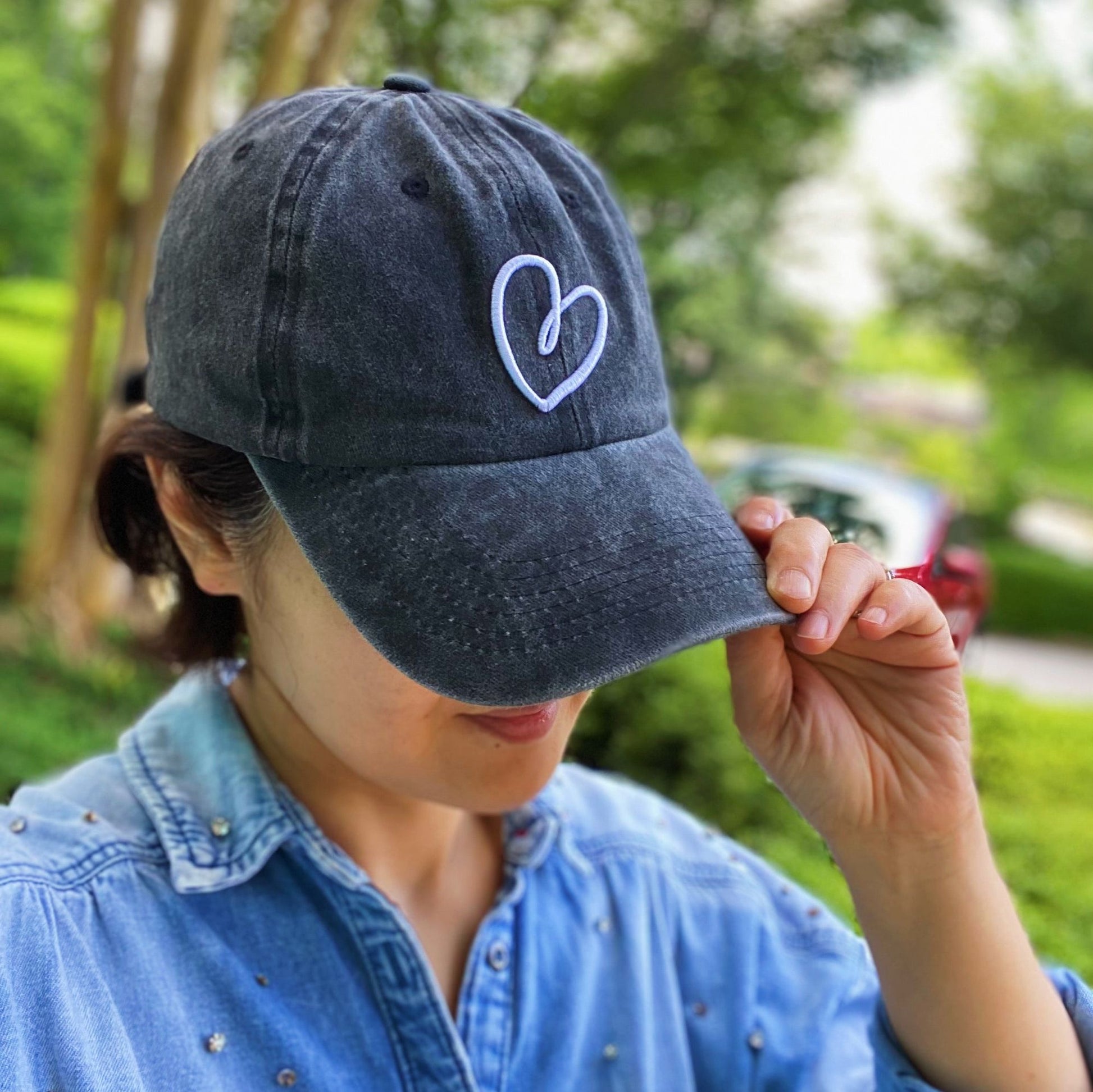 Stylish gray baseball cap with white logo, held by a person in a denim shirt, set against a lush green outdoor background.